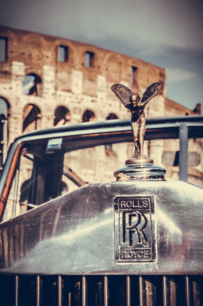 Il muso di una Rolls-Royce d'epoca con la celebre "Spirit of Ecstasy" in primo piano, sfondo suggestivo con il maestoso Colosseo di Roma